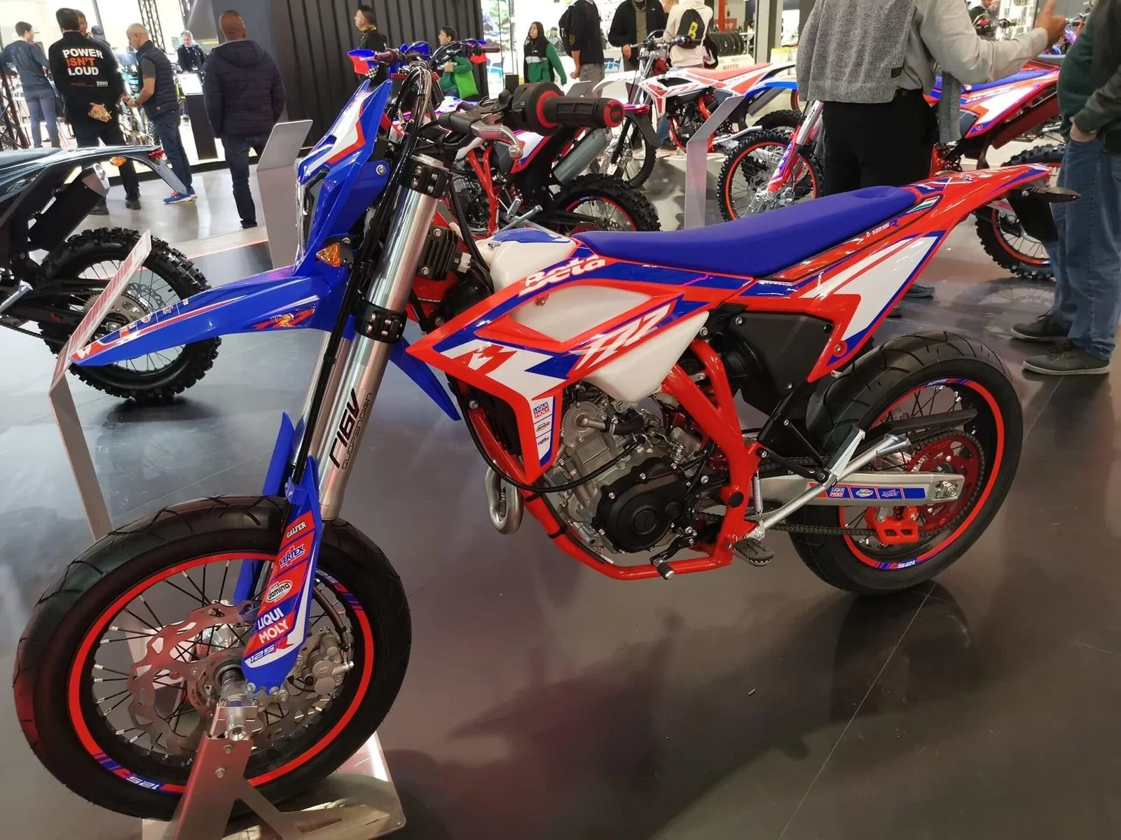 Moto supermotard rossa e blu esposta su un cavalletto in uno stand di EICMA